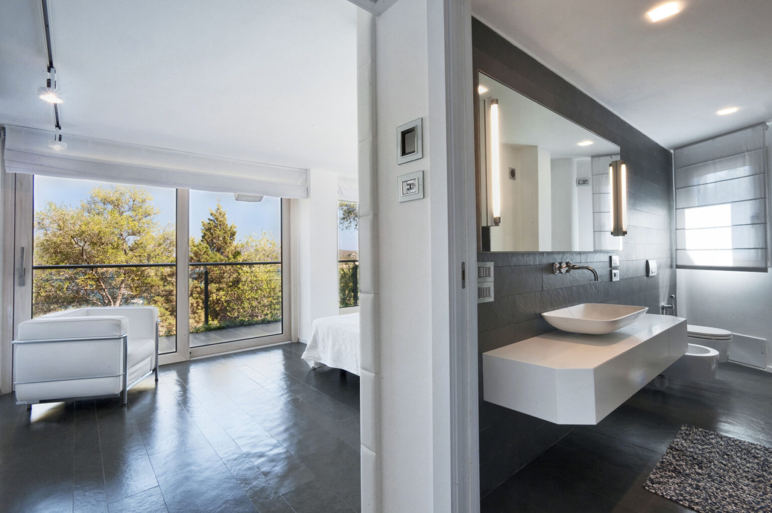 Modern interior of a stylish bedroom and bathroom with large windows showcasing greenery outside, featuring sleek furniture, minimalist decor, and contemporary lighting. | Only on SopranoVillas