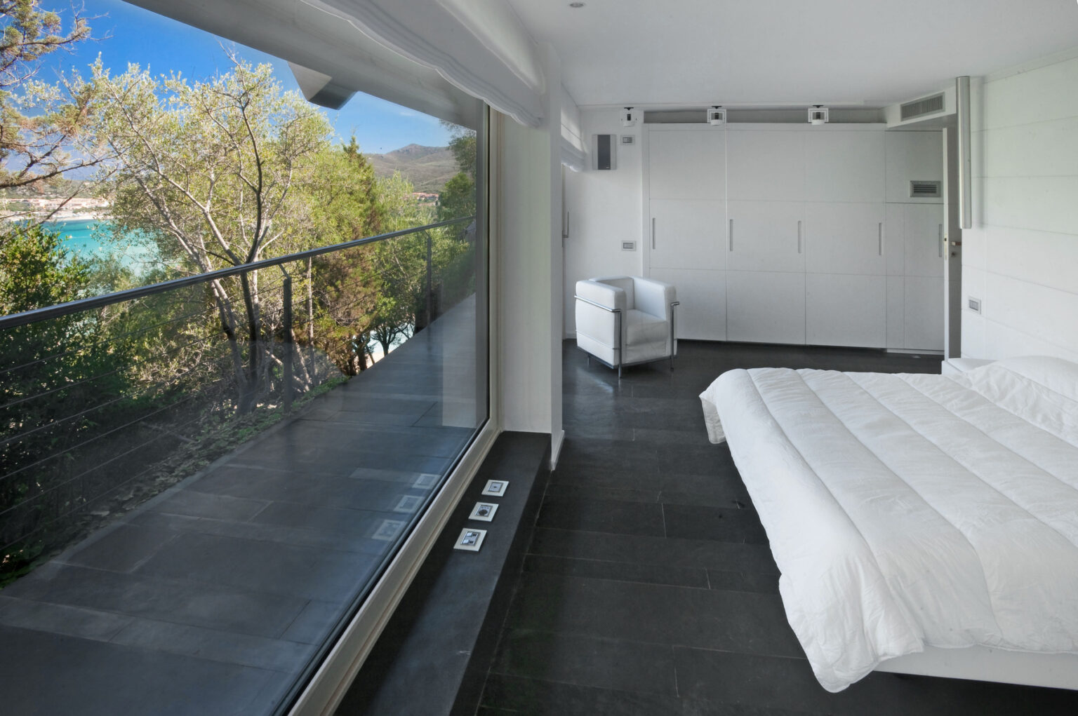 Modern bedroom with a large window showcasing a scenic view of trees and a beach. The minimalist design features a white bed, sleek furniture, and dark flooring, creating a serene atmosphere. | Only on SopranoVillas