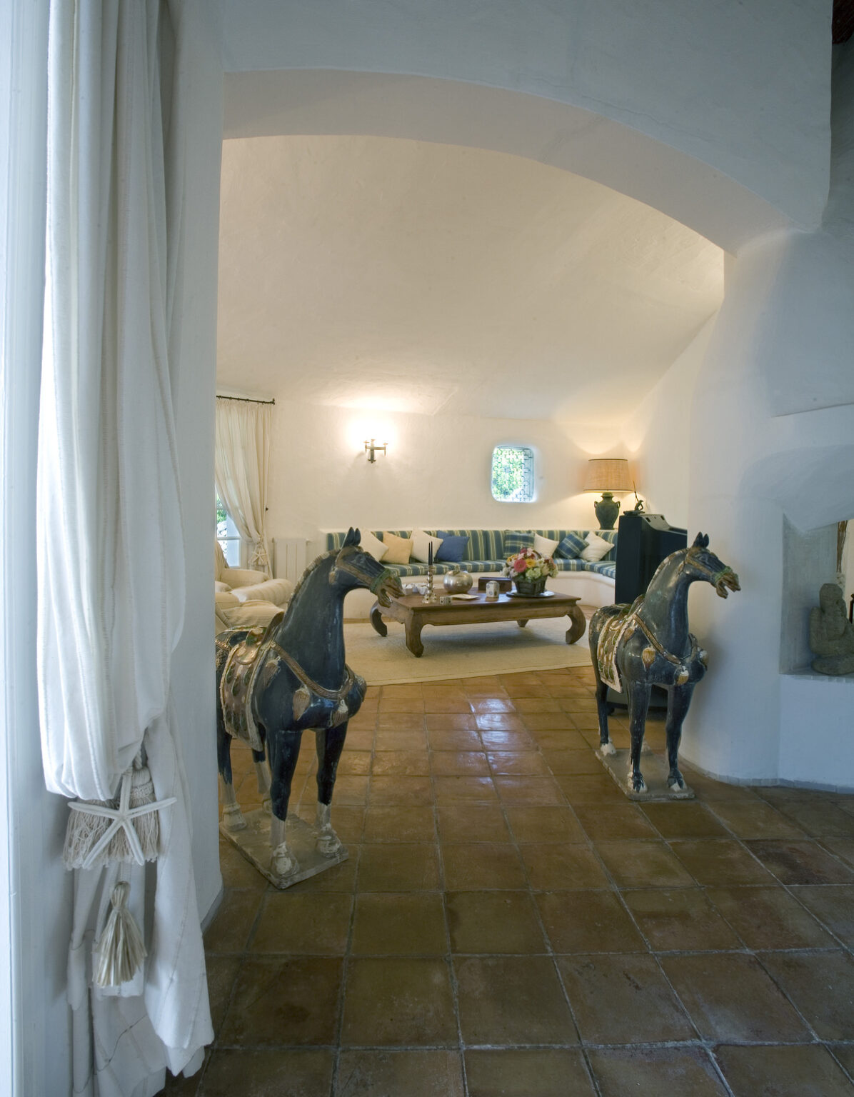 Interior view of a stylish living room featuring two decorative horse sculptures, a cozy couch with striped cushions, and a wooden coffee table. The space has a warm, inviting atmosphere with terracotta tiles and soft lighting, complemented by white walls and elegant curtains. | Only on SopranoVillas