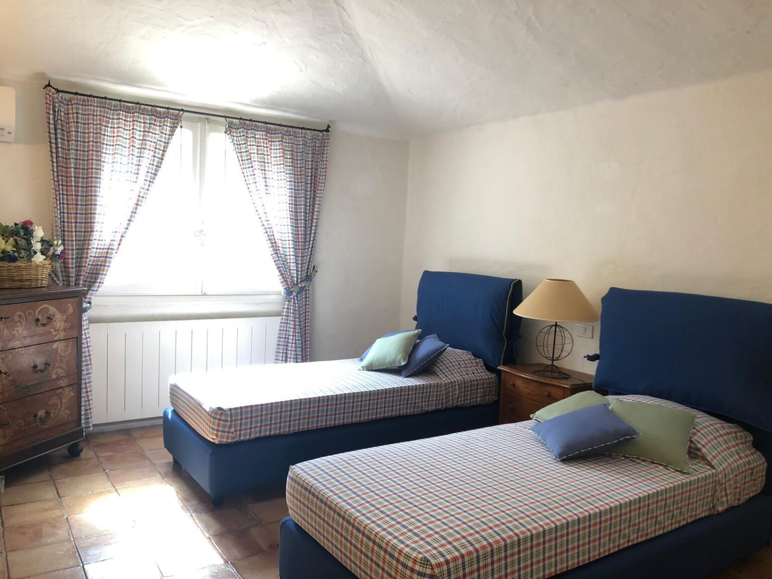Cozy bedroom featuring two twin beds with blue headboards, light-colored bedding, and decorative pillows, complemented by a wooden dresser and a lamp, with natural light streaming through a window adorned with checkered curtains. | Only on SopranoVillas