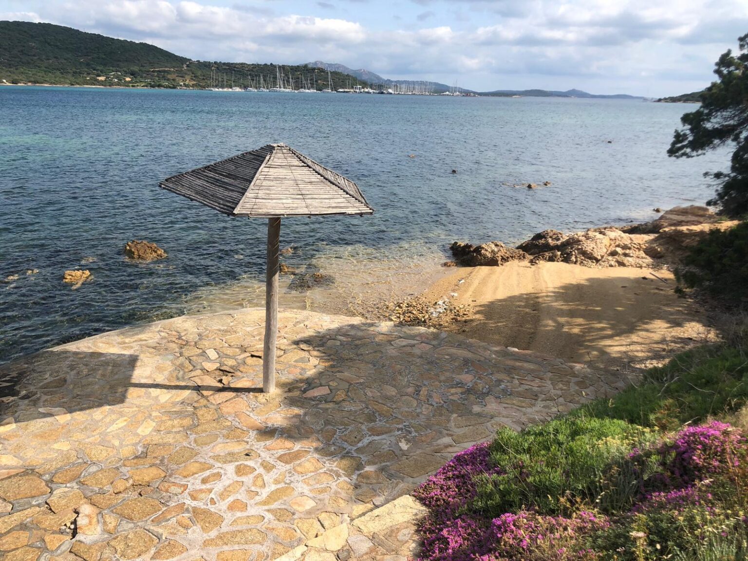 Scenic coastal view featuring a wooden beach umbrella on a stone pathway, with clear blue waters, rocky shores, and lush green hills in the background. Perfect for relaxation and enjoying nature. | Only on SopranoVillas
