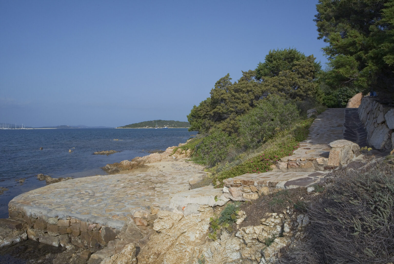 Coastal landscape featuring rocky shoreline, lush greenery, and calm blue waters under a clear sky. Ideal for nature lovers and photographers seeking serene outdoor locations. | Only on SopranoVillas