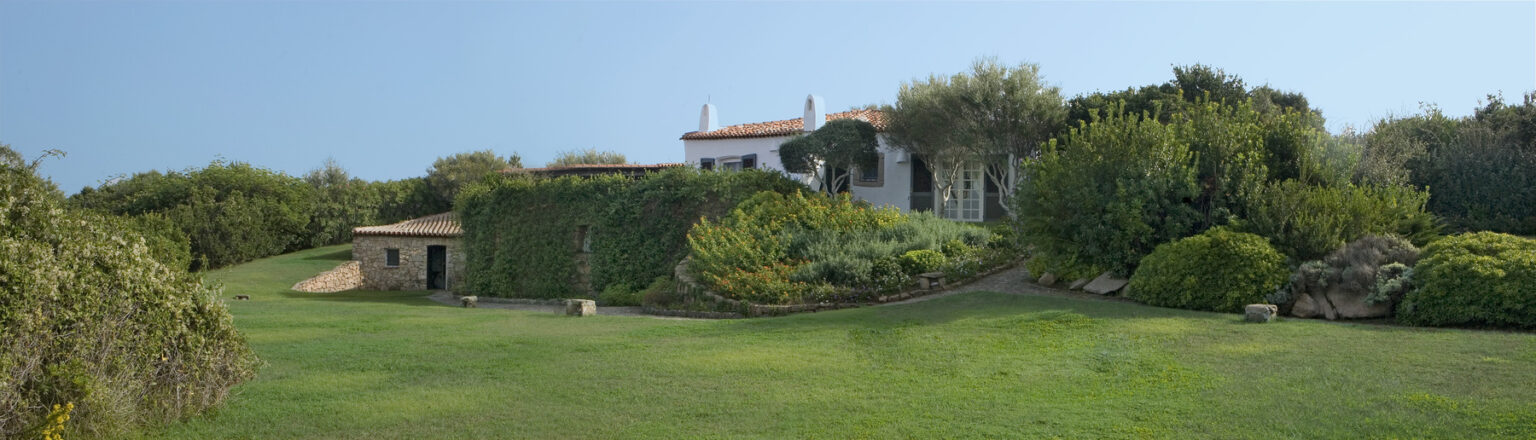 A picturesque view of a Mediterranean-style house surrounded by lush greenery and landscaped gardens, featuring a sloping lawn and decorative plants under a clear blue sky. | Only on SopranoVillas