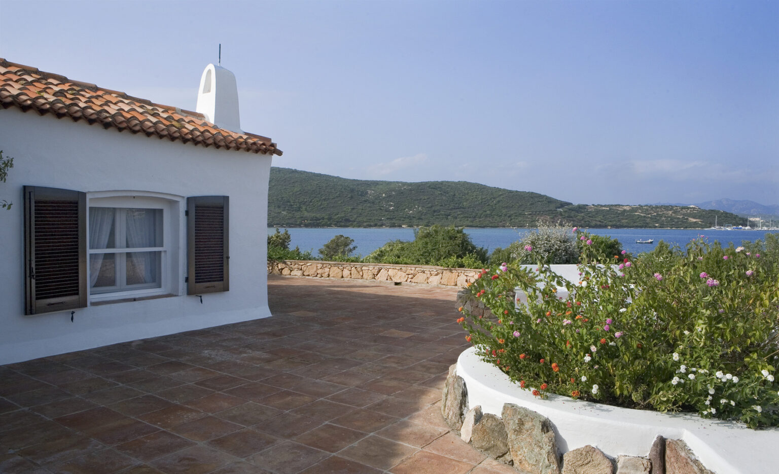 Charming white cottage with terracotta roof overlooking a serene bay, surrounded by colorful flowers and lush greenery. Ideal for a peaceful getaway in a coastal setting. | Only on SopranoVillas
