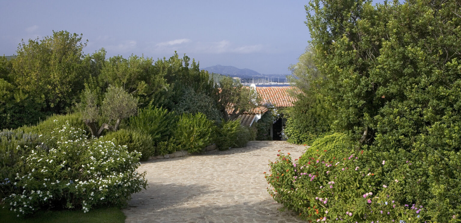 Scenic garden pathway leading to a charming house, surrounded by lush greenery and colorful flowers, with distant mountains and a marina visible in the background. | Only on SopranoVillas