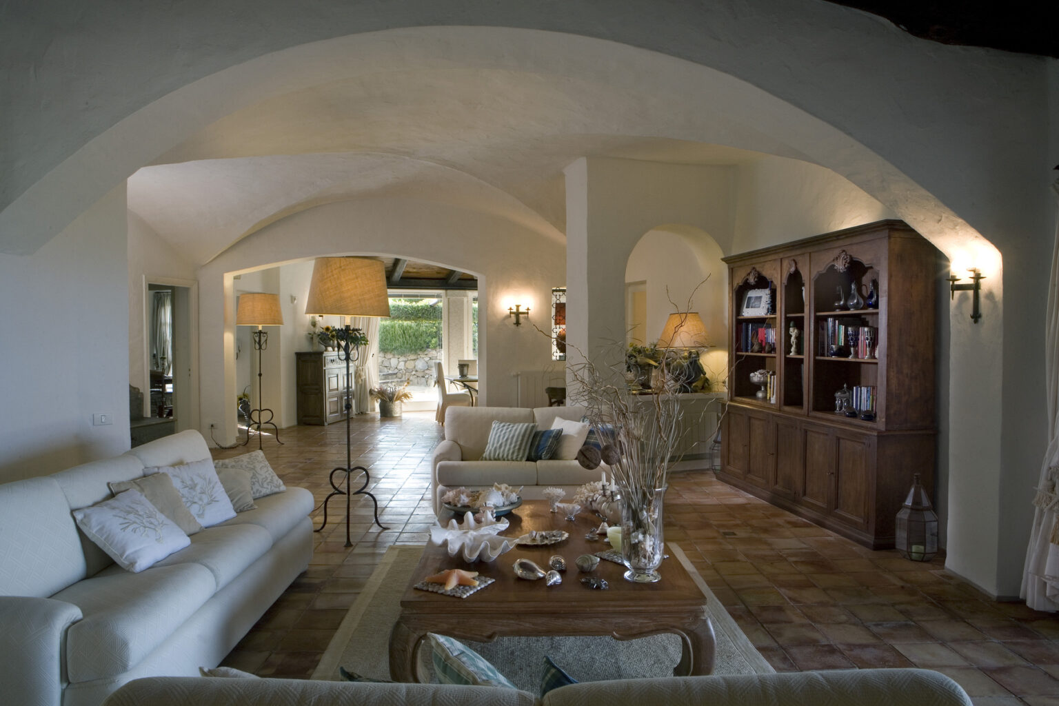 Spacious and elegantly furnished living room featuring arched ceilings, a comfortable white sofa, wooden coffee table, and a cozy reading nook, with natural light streaming through large windows. | Only on SopranoVillas