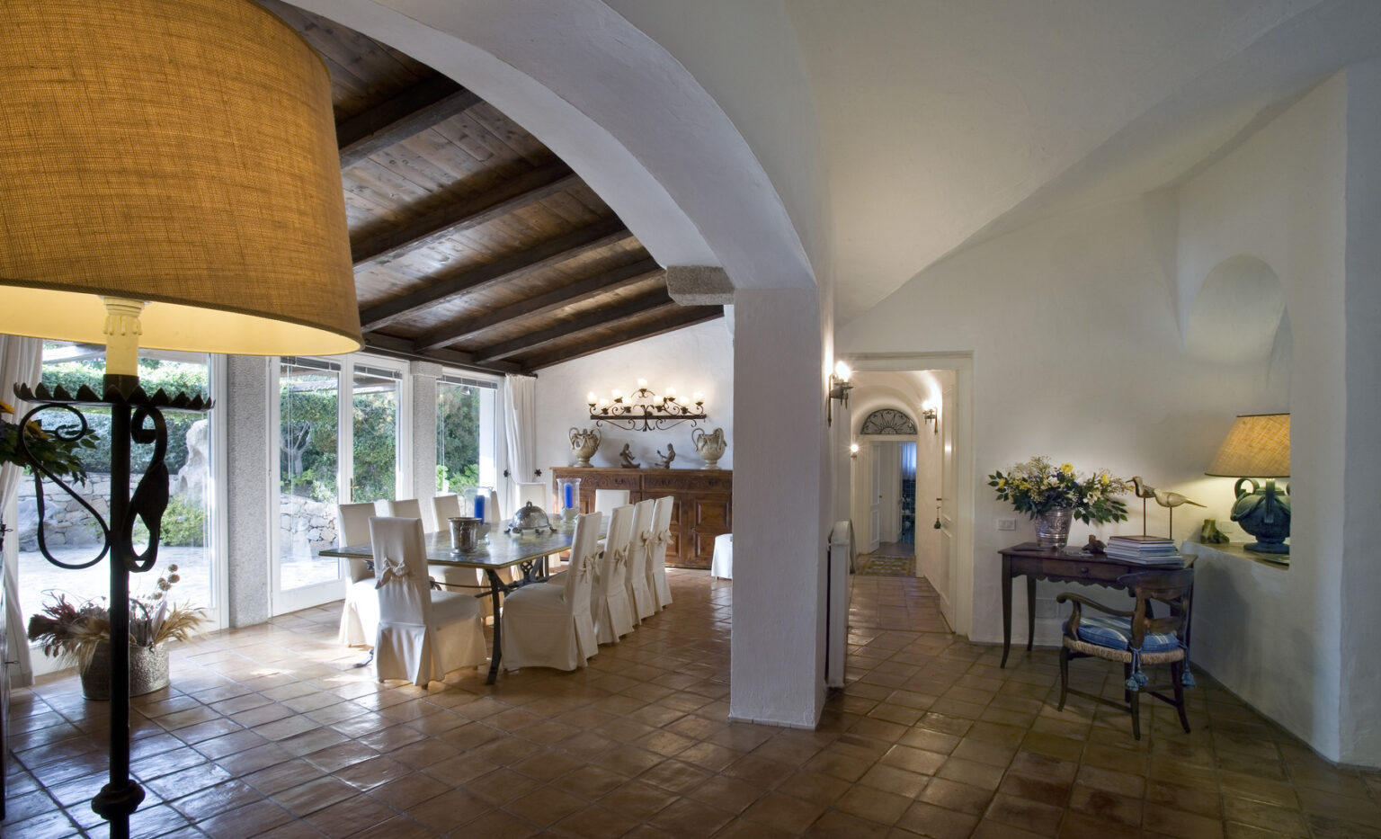 Spacious and elegant interior of a modern dining area featuring a large table, white chairs, and warm lighting, with arched ceilings and terracotta flooring, creating a welcoming atmosphere. | Only on SopranoVillas