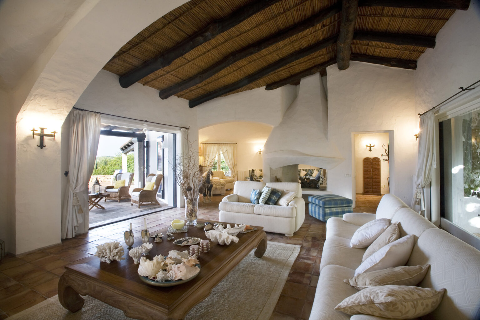 Spacious living room with high wooden beamed ceiling, featuring a cozy white sofa, stylish decor, and large windows opening to a scenic outdoor area. The room is elegantly designed with soft lighting and a warm color palette, creating a welcoming atmosphere. | Only on SopranoVillas