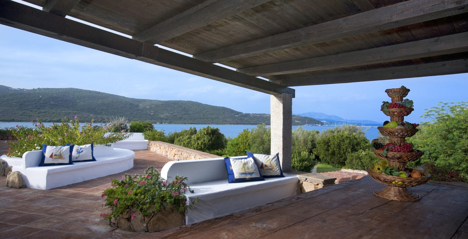 Scenic outdoor terrace with white seating adorned with decorative pillows, overlooking a tranquil bay and lush green hills. A multi-tiered fruit basket adds a touch of elegance to the setting, ideal for relaxation and enjoying nature. | Only on SopranoVillas
