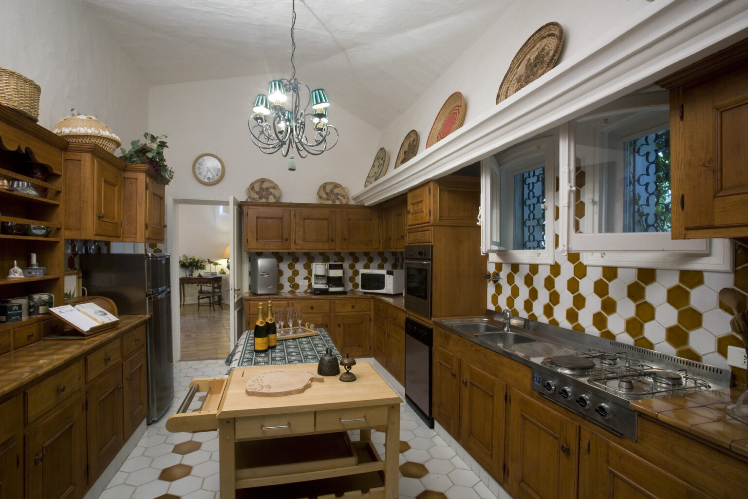 Cozy rustic kitchen featuring wooden cabinets, a central island with a cutting board, and modern appliances. The walls are adorned with decorative plates and honeycomb-patterned tiles, illuminated by a stylish chandelier. | Only on SopranoVillas