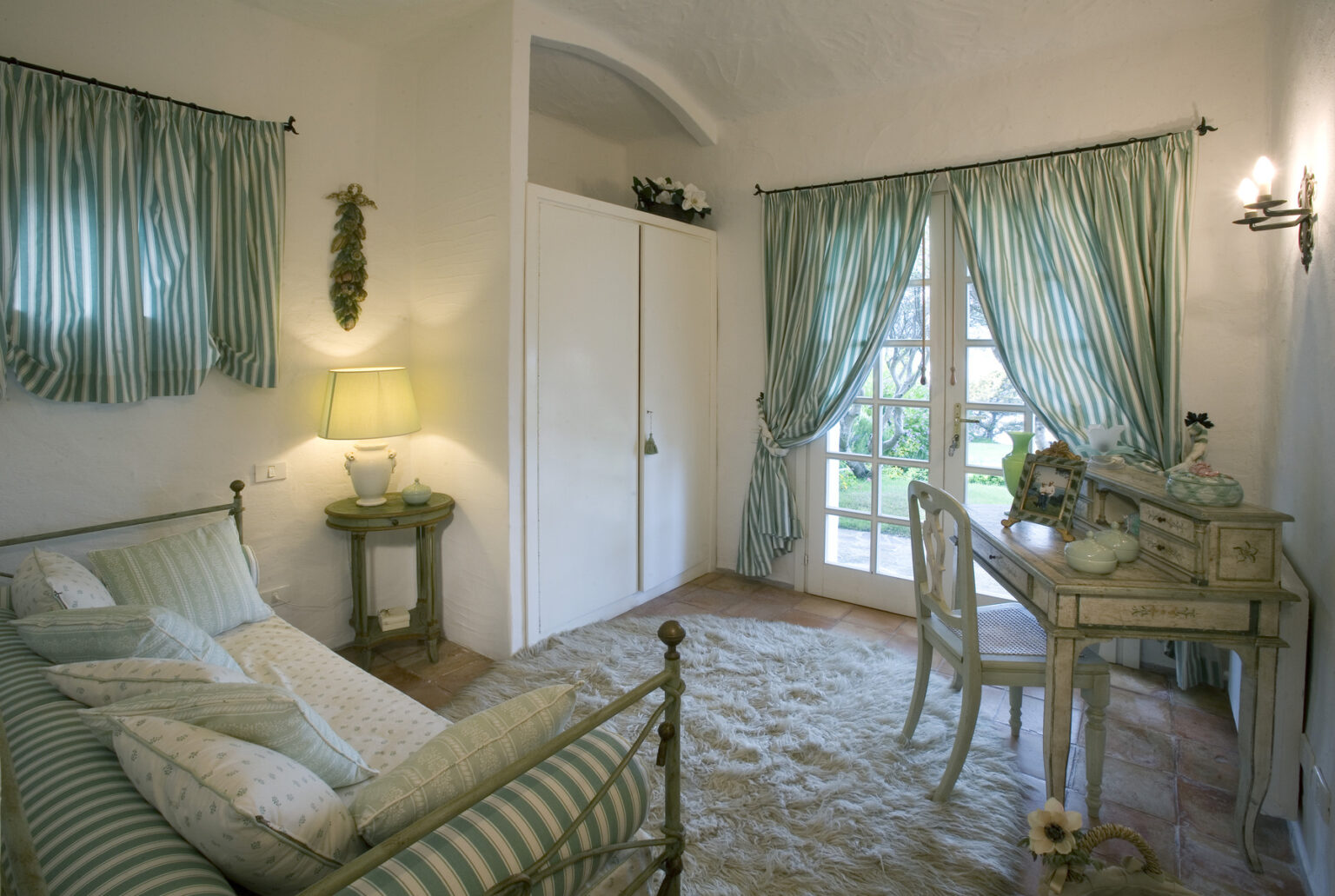 Cozy bedroom interior featuring a light green and white color scheme, a brass bed, a vintage writing desk, and elegant curtains. The space is well-lit with natural light streaming through large windows, creating a serene and inviting atmosphere. | Only on SopranoVillas