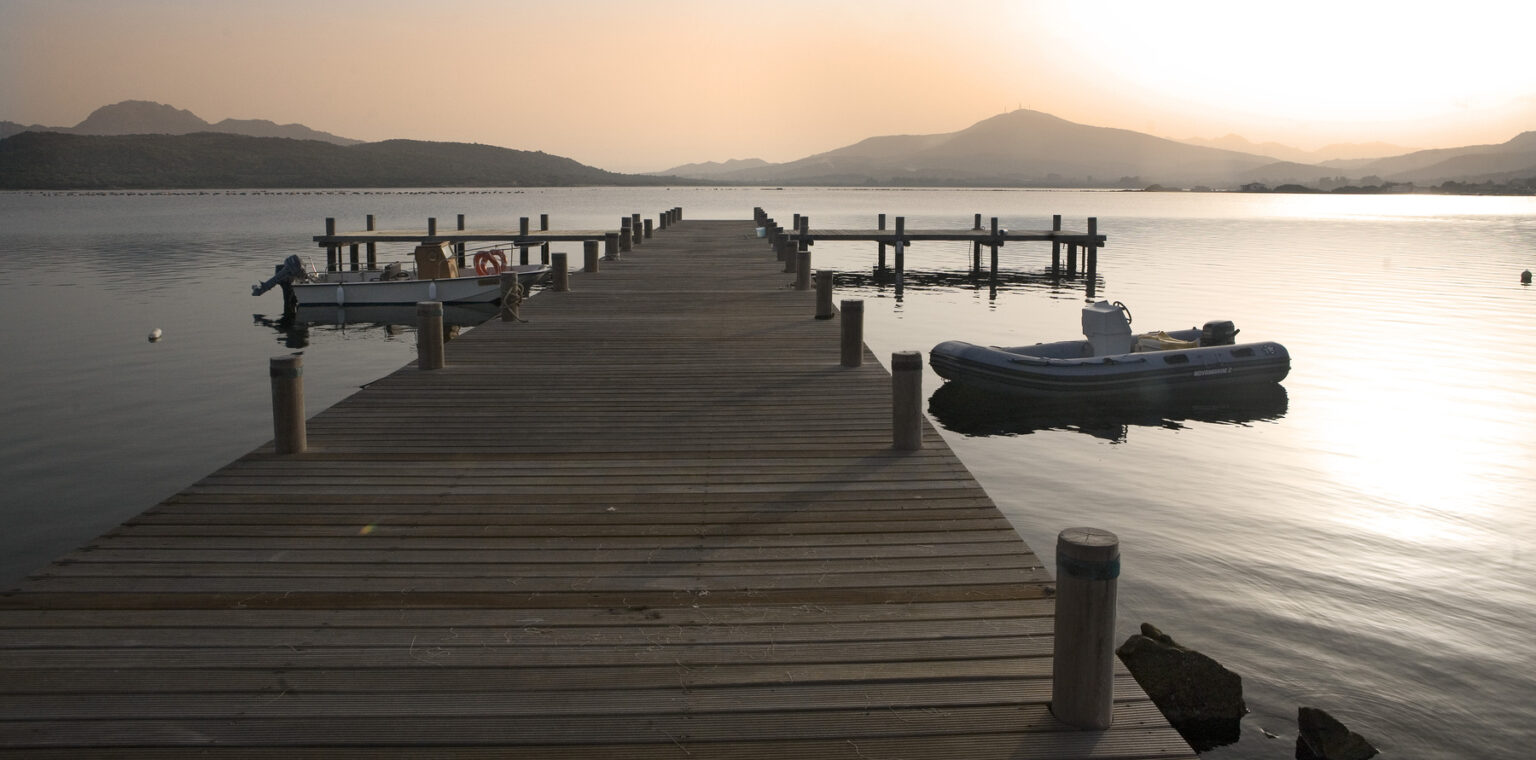 Wooden pier extending over calm water at sunset, with small boats moored alongside and mountains in the background. | Only on SopranoVillas