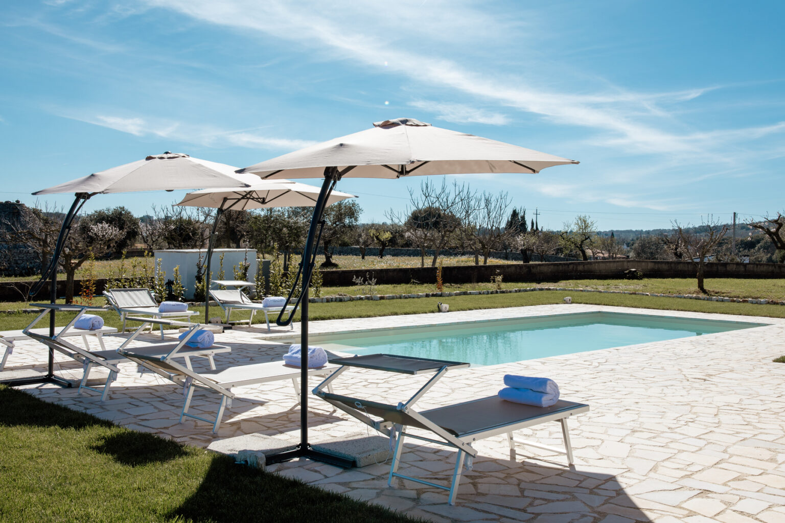 A serene outdoor pool area featuring a clear blue swimming pool, comfortable lounge chairs, and large umbrellas, surrounded by lush greenery and a clear blue sky. Ideal for relaxation and leisure. | Only on SopranoVillas