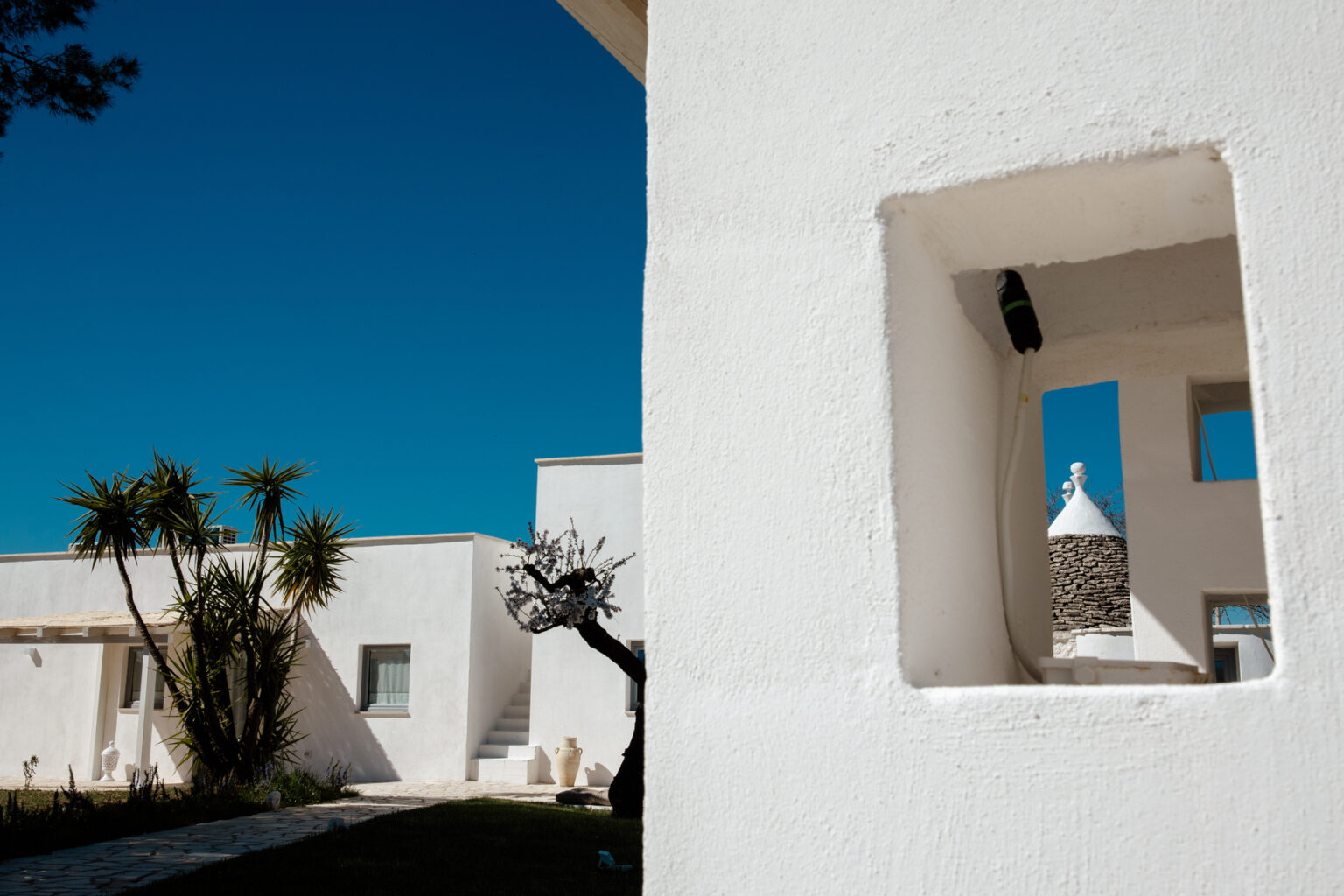 Modern architectural design featuring a white building with a window frame, surrounded by lush greenery and a clear blue sky. The image highlights unique structural elements and a serene outdoor space. | Only on SopranoVillas