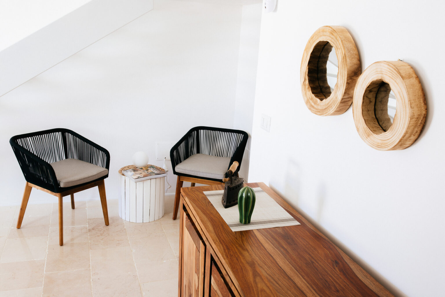 Modern living room interior featuring two black wicker chairs with cushions, a wooden side table, and decorative wall art made of natural materials. The space is bright and minimalistic, emphasizing a cozy and stylish atmosphere. | Only on SopranoVillas