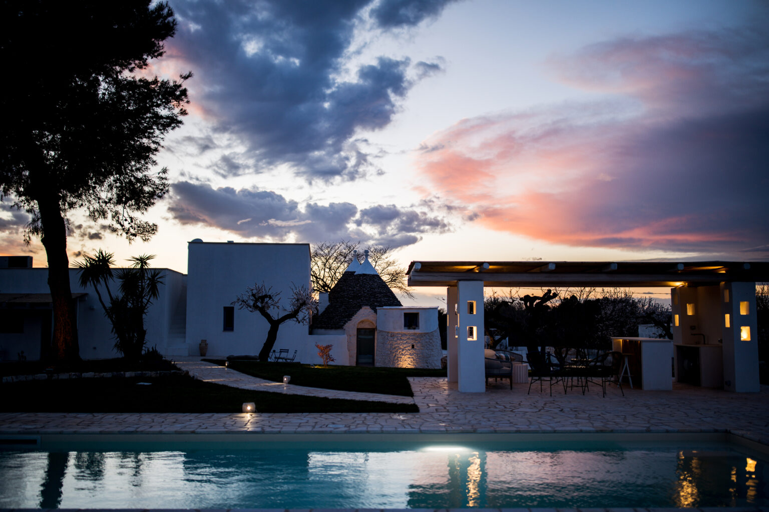 Modern villa with a swimming pool at sunset, featuring white architecture, landscaped gardens, and dramatic cloud formations in the sky. | Only on SopranoVillas