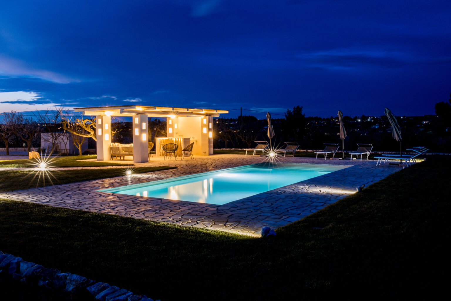 Modern outdoor pool area illuminated at night, featuring a pergola with string lights, lounge chairs, and a landscaped garden. Ideal for relaxing and entertaining in a tranquil setting. | Only on SopranoVillas