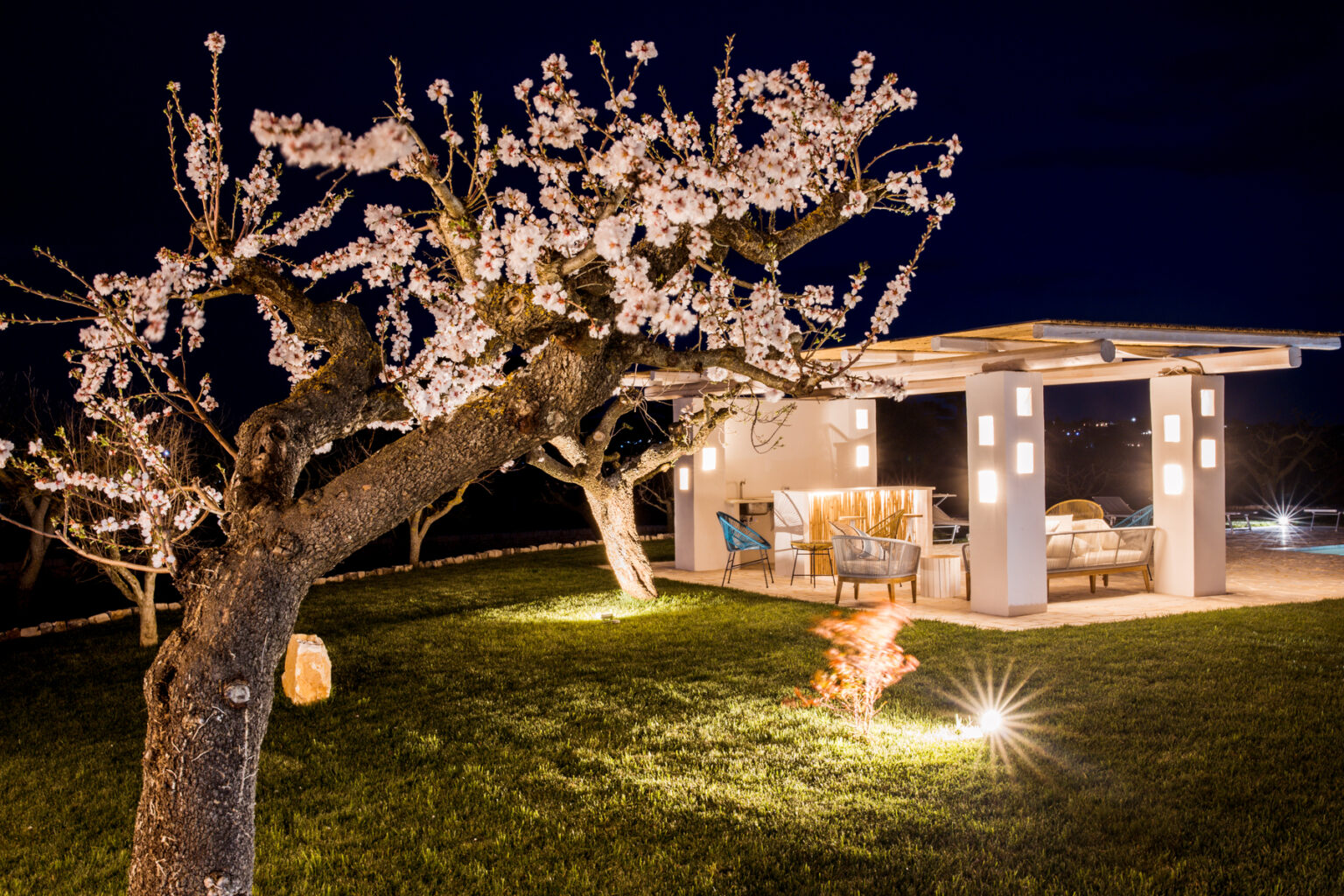 Cozy outdoor seating area illuminated at night, surrounded by a blooming cherry blossom tree and garden lights, creating a serene atmosphere for relaxation. | Only on SopranoVillas
