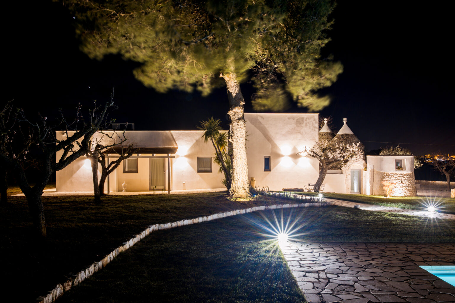 Charming illuminated villa at night featuring traditional trullo architecture, surrounded by landscaped gardens and trees, with a stone pathway and pool area. | Only on SopranoVillas