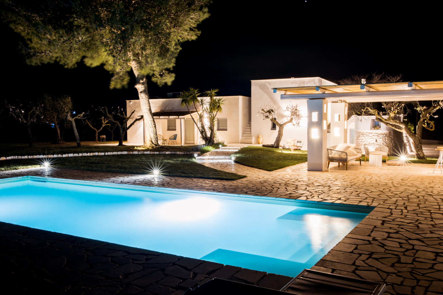 Modern villa with a glowing pool and landscaped garden at night, featuring outdoor seating and illuminated trees. | Only on SopranoVillas