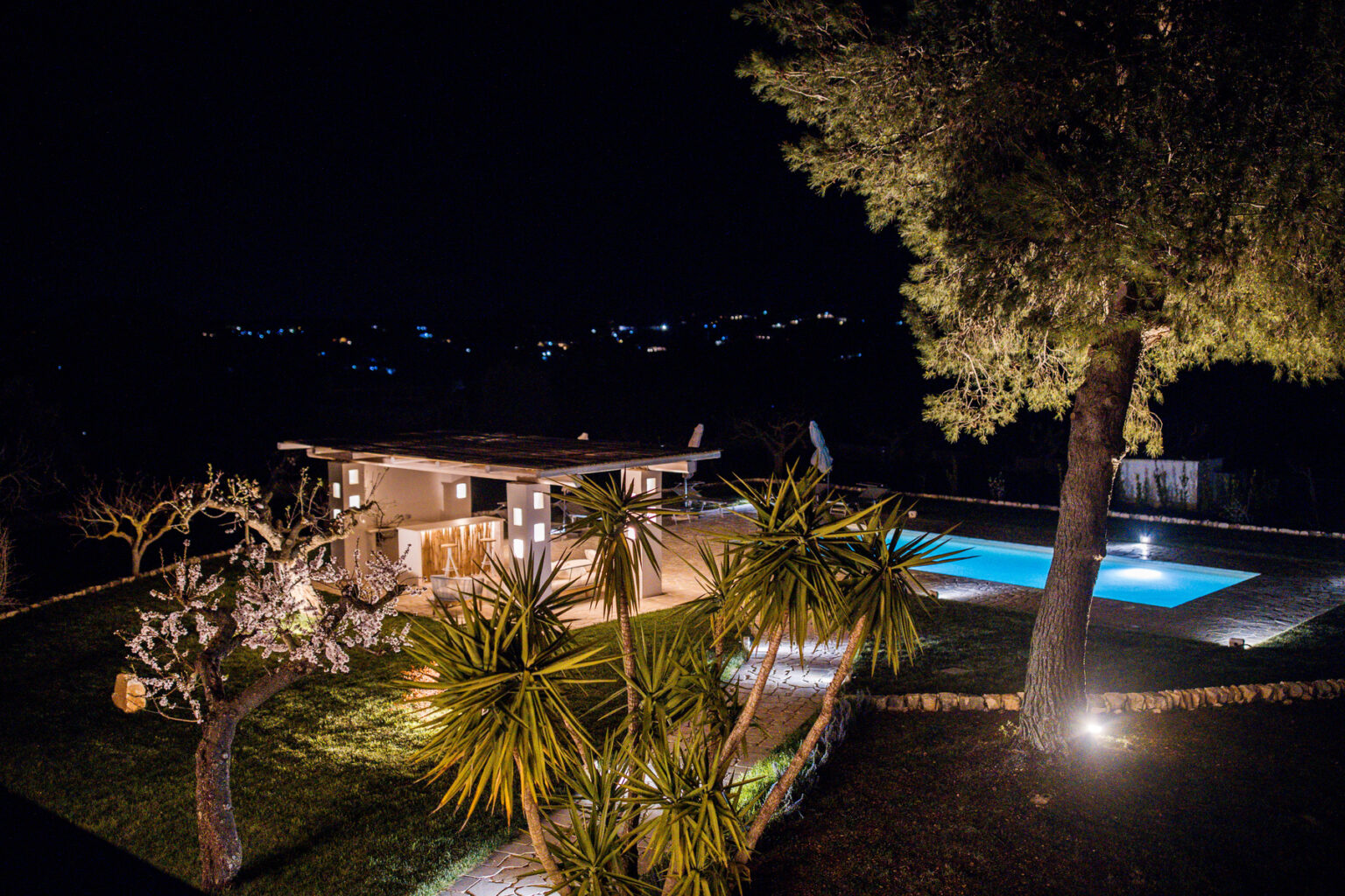 Modern villa at night with illuminated landscaping and a swimming pool, surrounded by lush greenery and trees. | Only on SopranoVillas