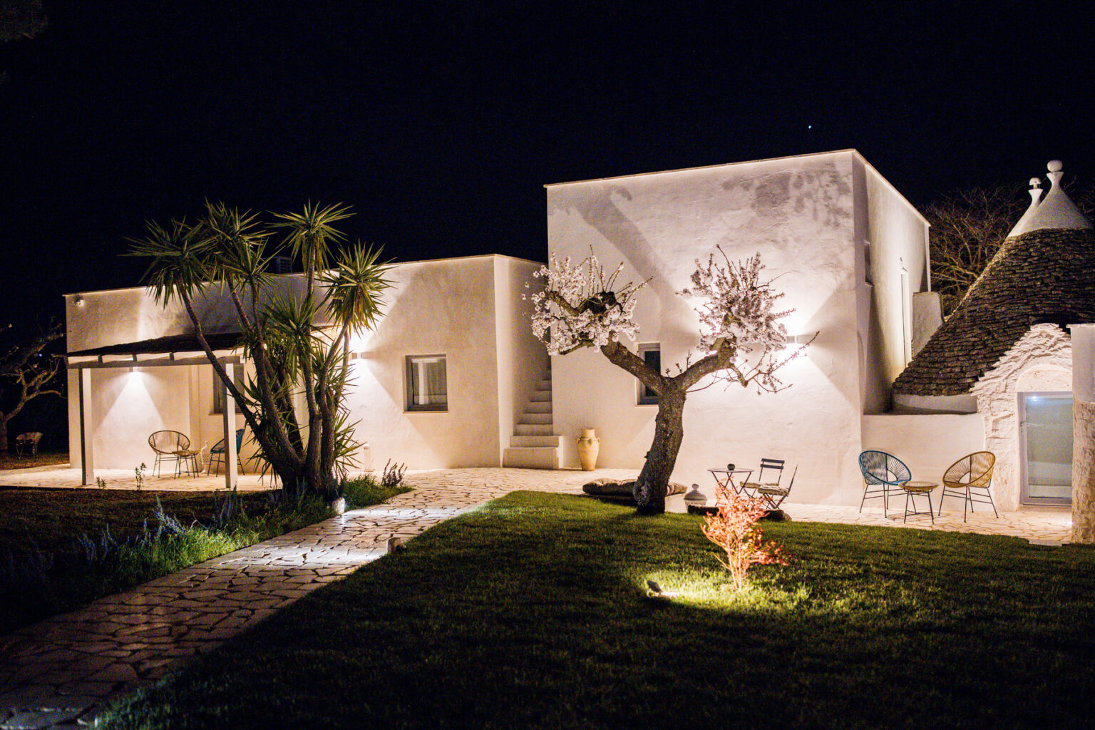 Charming white villa with illuminated exterior at night, featuring a traditional trullo roof, landscaped garden, and outdoor seating area. | Only on SopranoVillas