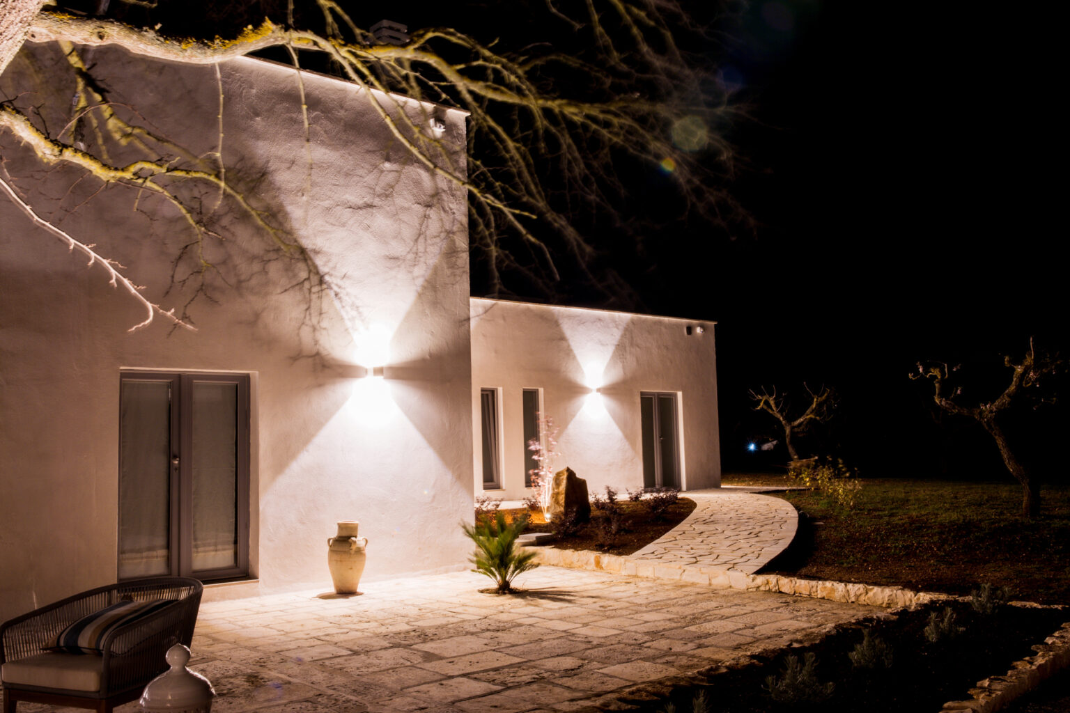 Modern house exterior illuminated at night with soft lighting, showcasing a stone pathway, decorative plants, and an elegant seating area. The minimalist design features smooth white walls and large windows, set against a dark, tranquil landscape. | Only on SopranoVillas