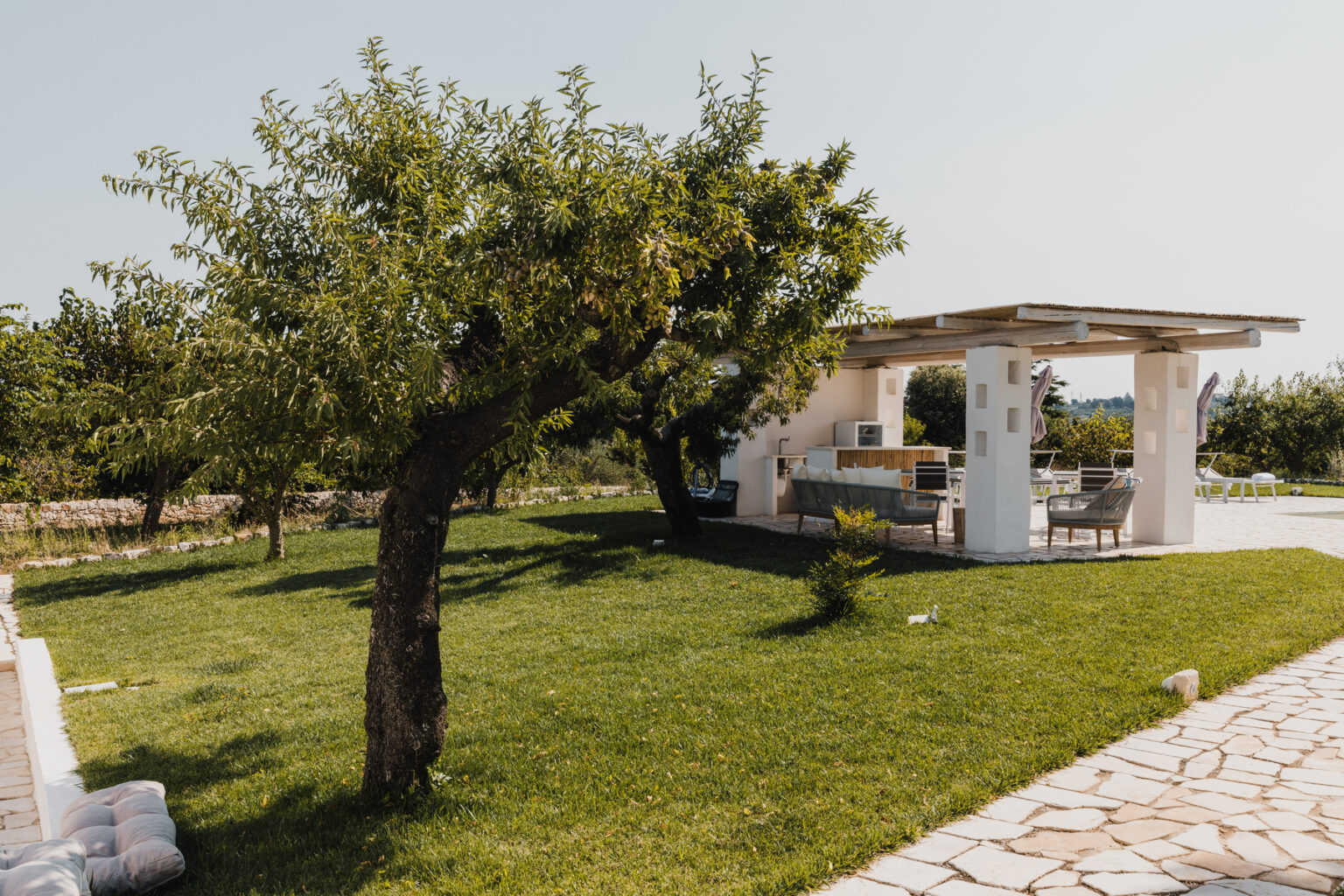 Scenic view of a landscaped garden featuring a lush green lawn, a fruit tree, and a modern outdoor kitchen area with seating under a shaded patio. | Only on SopranoVillas