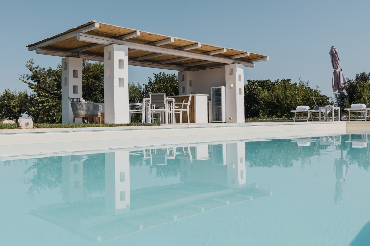 Modern outdoor pool area featuring a stylish pergola, dining set, and lounge chairs surrounded by greenery, reflecting in the clear water. Ideal for relaxation and entertaining. | Only on SopranoVillas