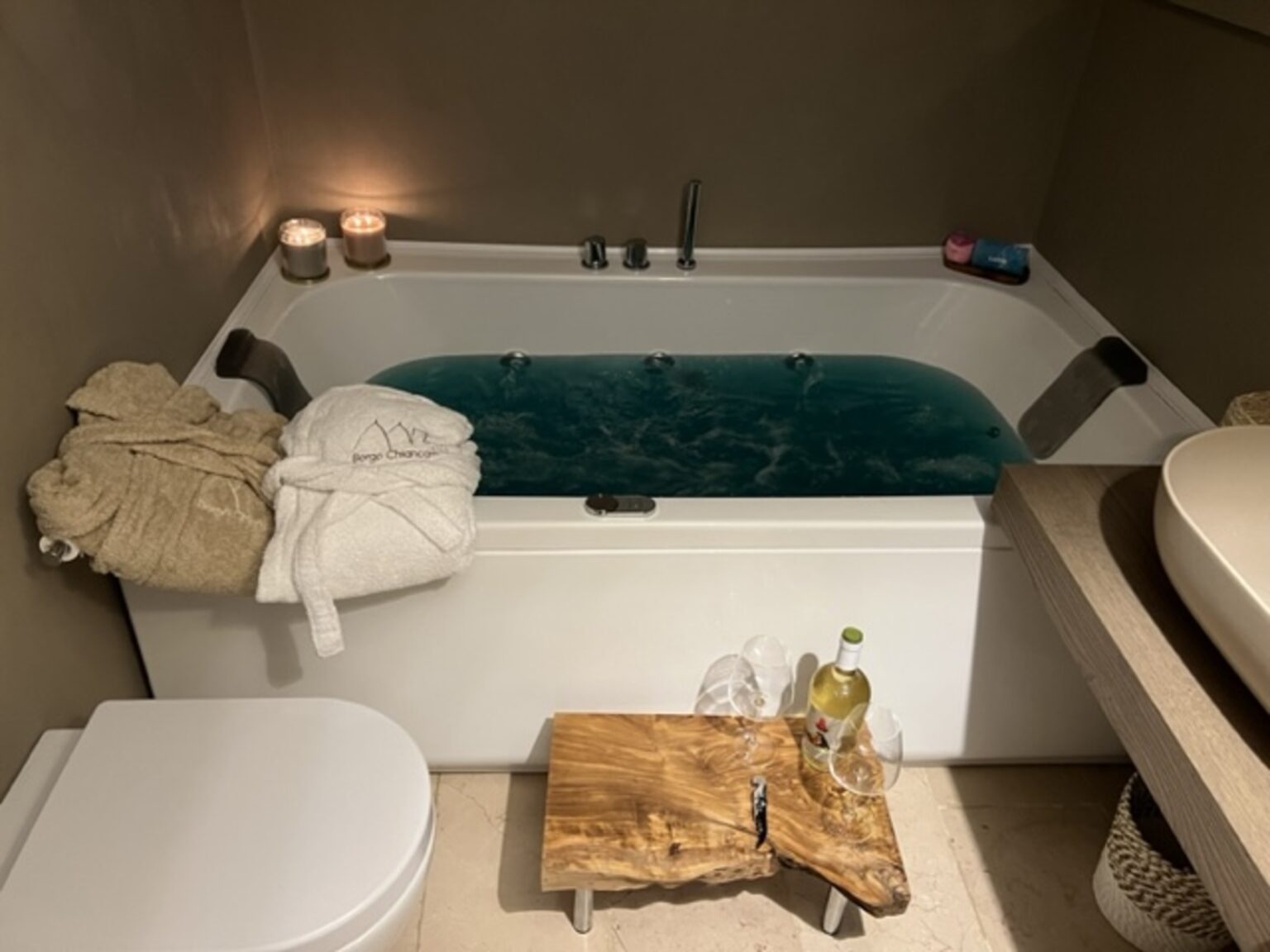Luxurious bathroom featuring a modern jacuzzi tub with blue water, surrounded by soft towels, candles, and a wooden accent table holding wine glasses and a bottle of white wine, creating a relaxing spa-like atmosphere. | Only on SopranoVillas