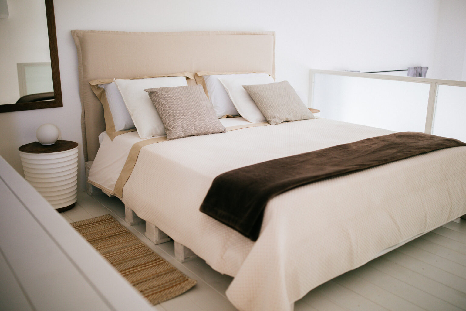 Cozy bedroom featuring a beige upholstered bed with multiple decorative pillows, a soft brown throw blanket, and a stylish nightstand with a round lamp. The room has a minimalist design with white walls and wooden accents, creating a serene atmosphere. | Only on SopranoVillas