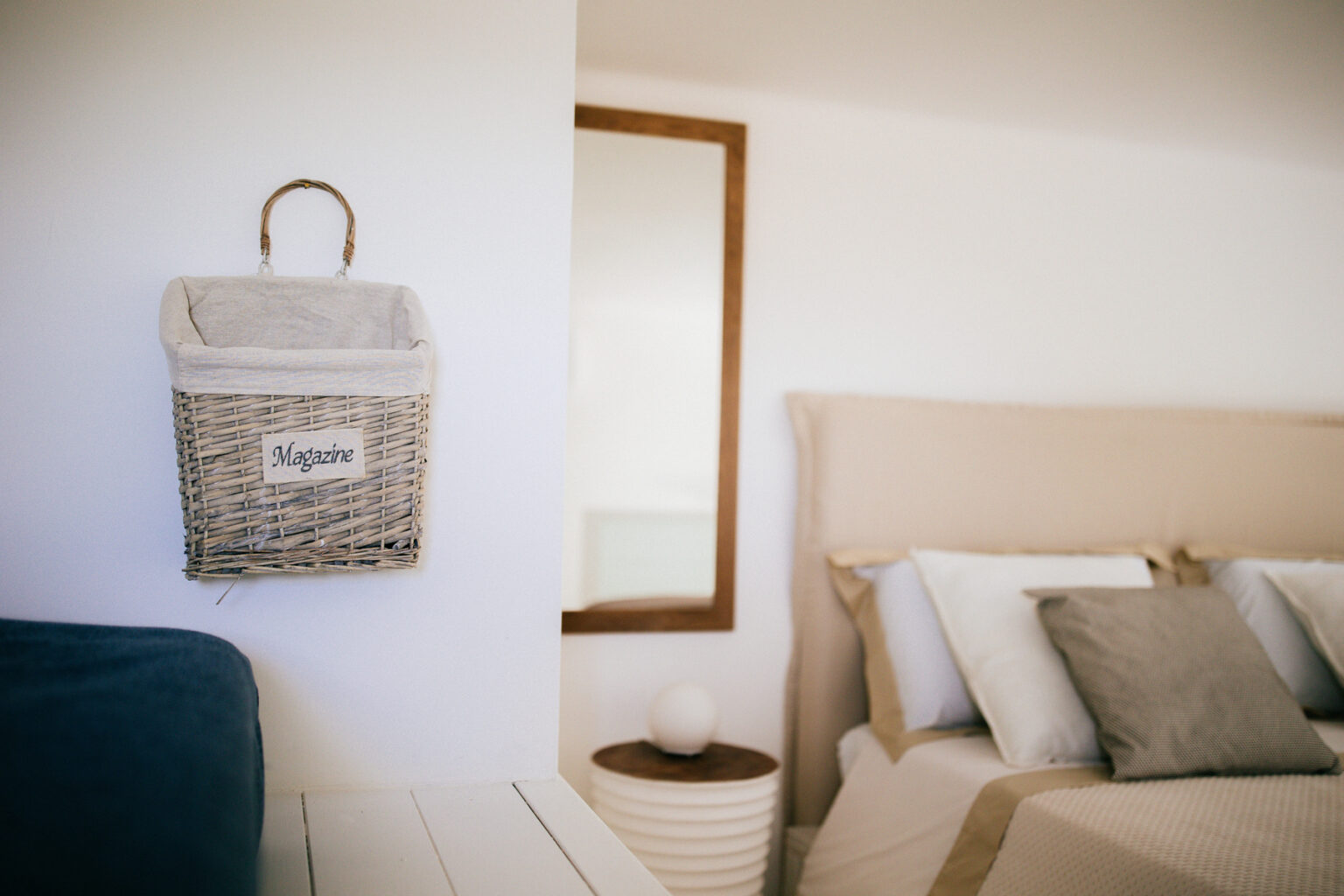 Cozy bedroom interior featuring a neatly made bed with decorative pillows, a stylish round side table, and a woven basket hanging on the wall labeled 'Bags.' Natural light enhances the serene atmosphere. | Only on SopranoVillas