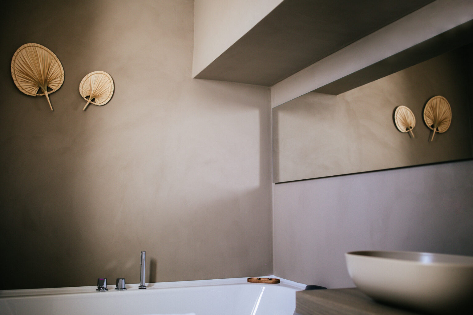 Modern bathroom interior featuring a minimalist design, gray walls, decorative wall art resembling palm leaves, a spacious bathtub, and a large mirror reflecting natural light. | Only on SopranoVillas