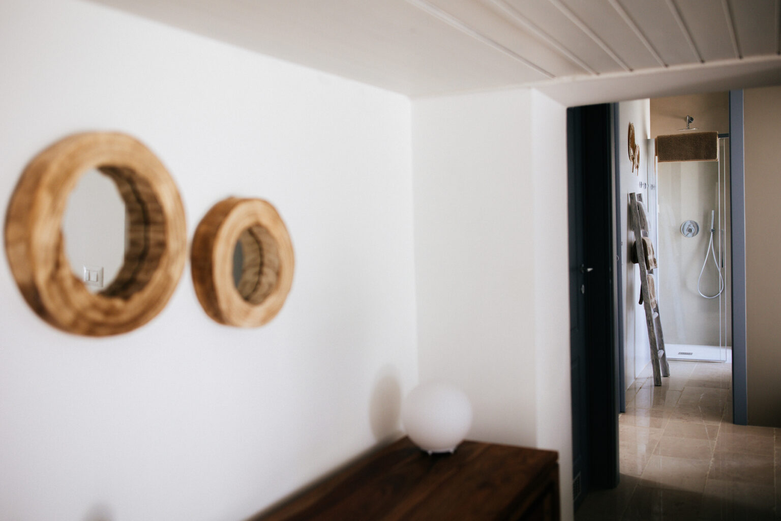 Interior view of a modern hallway featuring two round wooden mirrors on a white wall, leading to a bright bathroom with a shower in the background. The space showcases a minimalist design with natural elements. | Only on SopranoVillas