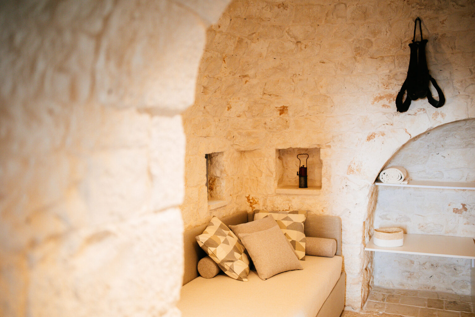 Cozy interior of a rustic room featuring stone walls, a minimalist sofa adorned with decorative pillows, and a small shelf displaying rolled towels and a decorative basket. A vintage lantern is placed on a ledge, enhancing the warm and inviting atmosphere. | Only on SopranoVillas