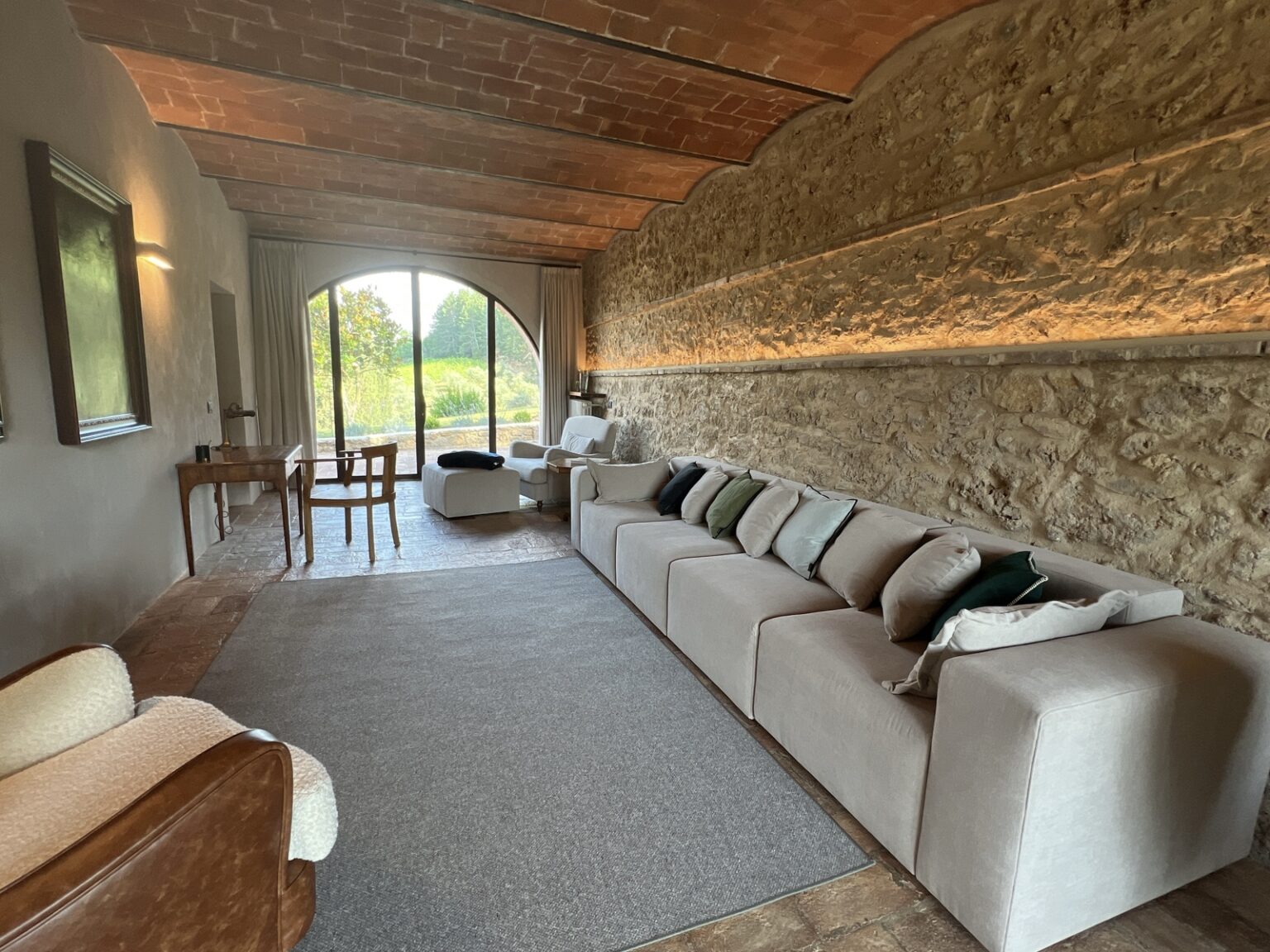 Modern living room with a stone wall, large windows, and a cozy sofa adorned with pillows, featuring wooden furniture and warm lighting for a welcoming atmosphere. | Only on SopranoVillas