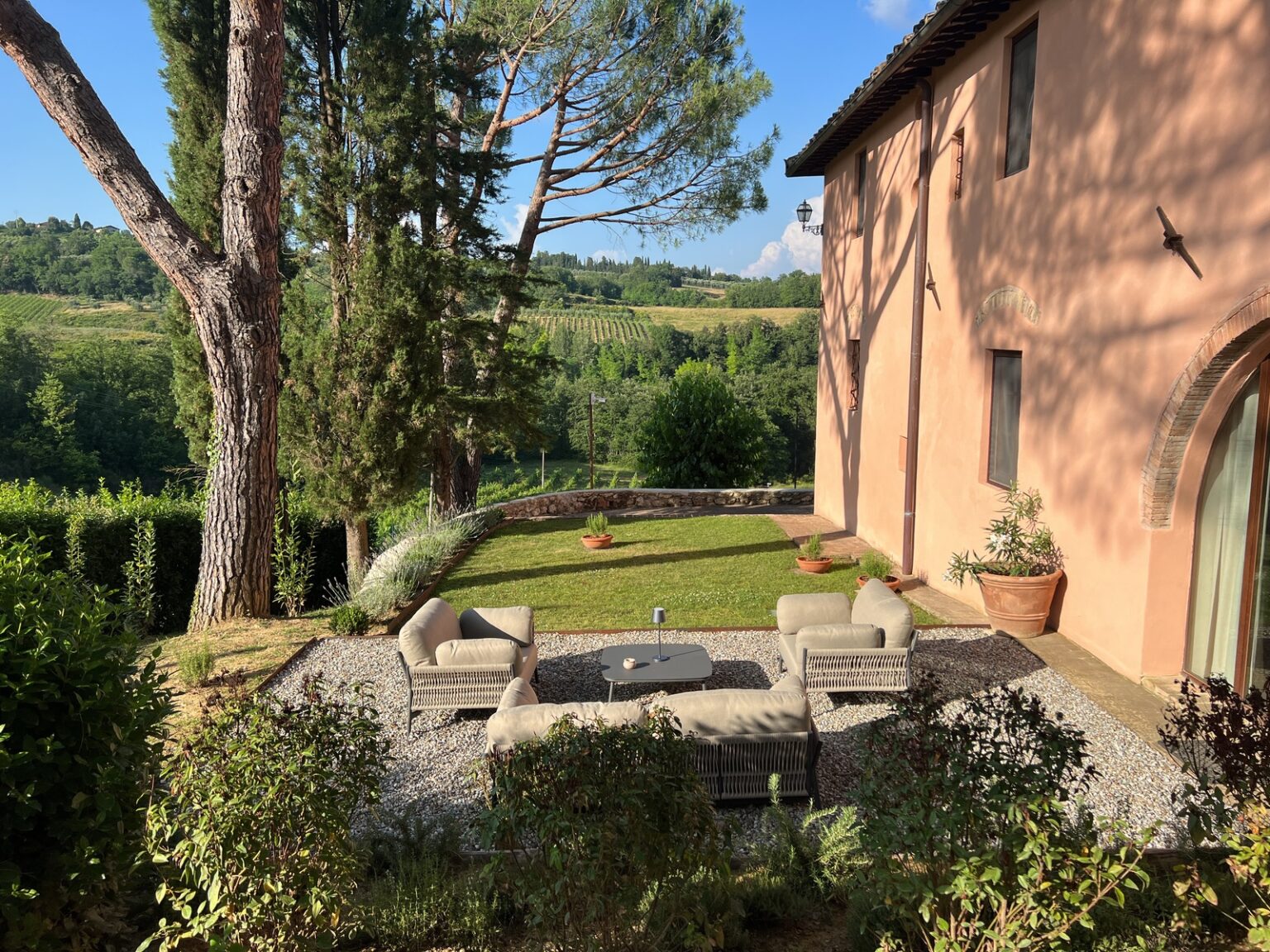 Cozy outdoor seating area with wicker furniture on a lawn, surrounded by lush greenery and trees, adjacent to a rustic building in a scenic landscape. | Only on SopranoVillas
