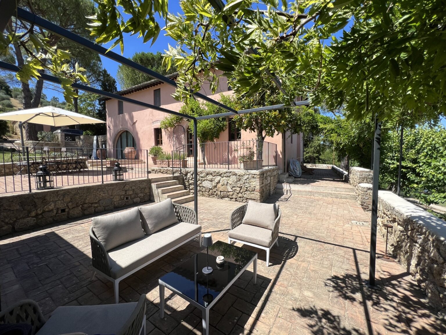 Outdoor seating area with modern furniture, surrounded by lush greenery and a charming pink villa in the background, perfect for relaxing or entertaining. | Only on SopranoVillas