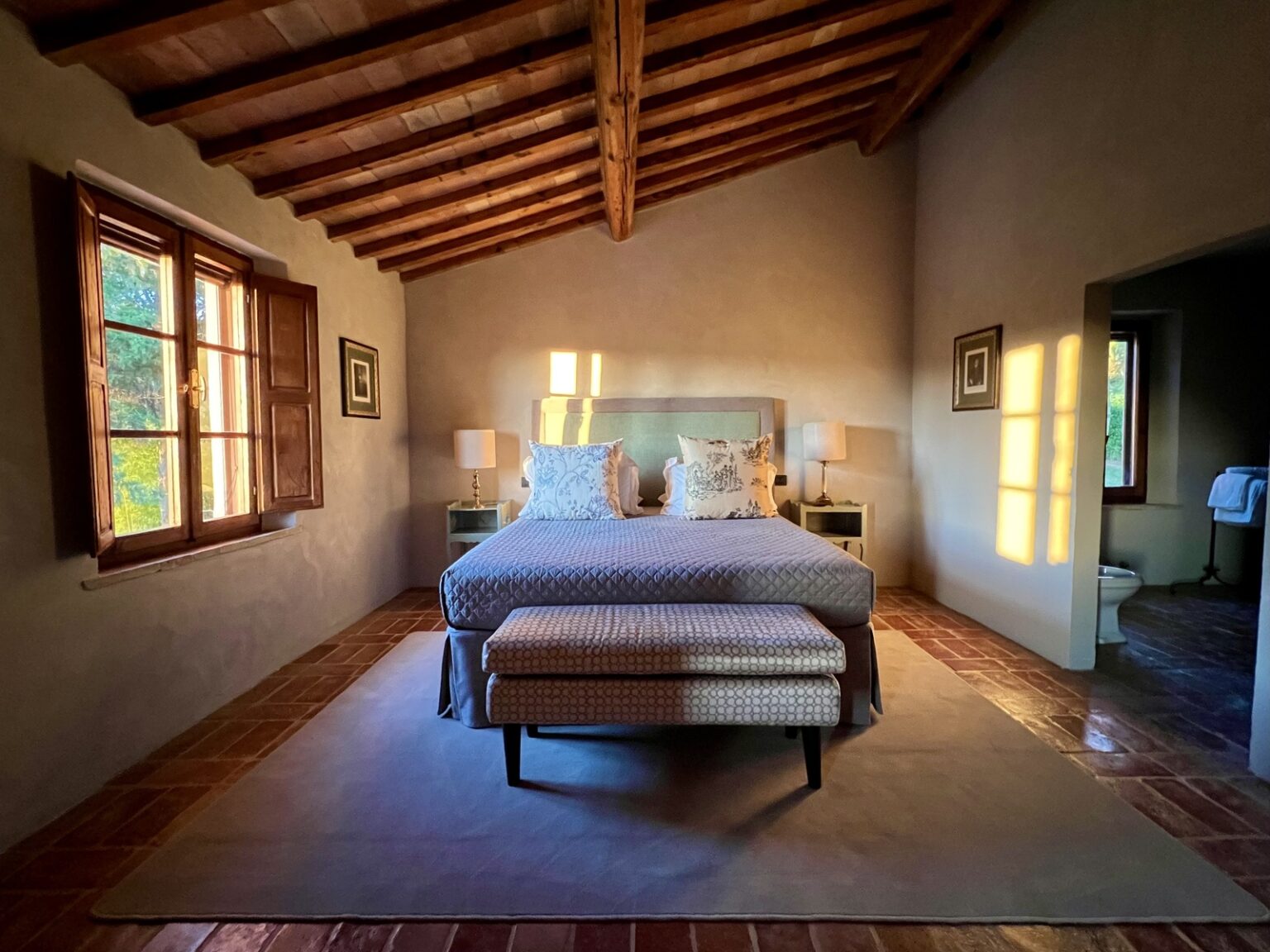 Cozy bedroom featuring a king-sized bed with decorative pillows, wooden beams on the ceiling, and large windows allowing natural light to fill the space. The room includes stylish bedside lamps and a comfortable bench at the foot of the bed, creating a warm and inviting atmosphere. | Only on SopranoVillas