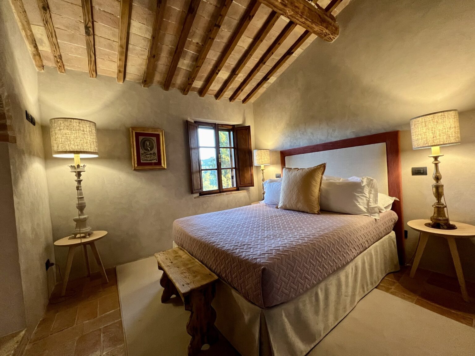 Cozy bedroom with a queen-sized bed, soft bedding, and decorative pillows, featuring rustic wooden beams on the ceiling. Two stylish bedside lamps provide warm lighting, and a vintage framed picture adds character to the wall. A wooden bench sits at the foot of the bed, enhancing the inviting atmosphere. | Only on SopranoVillas