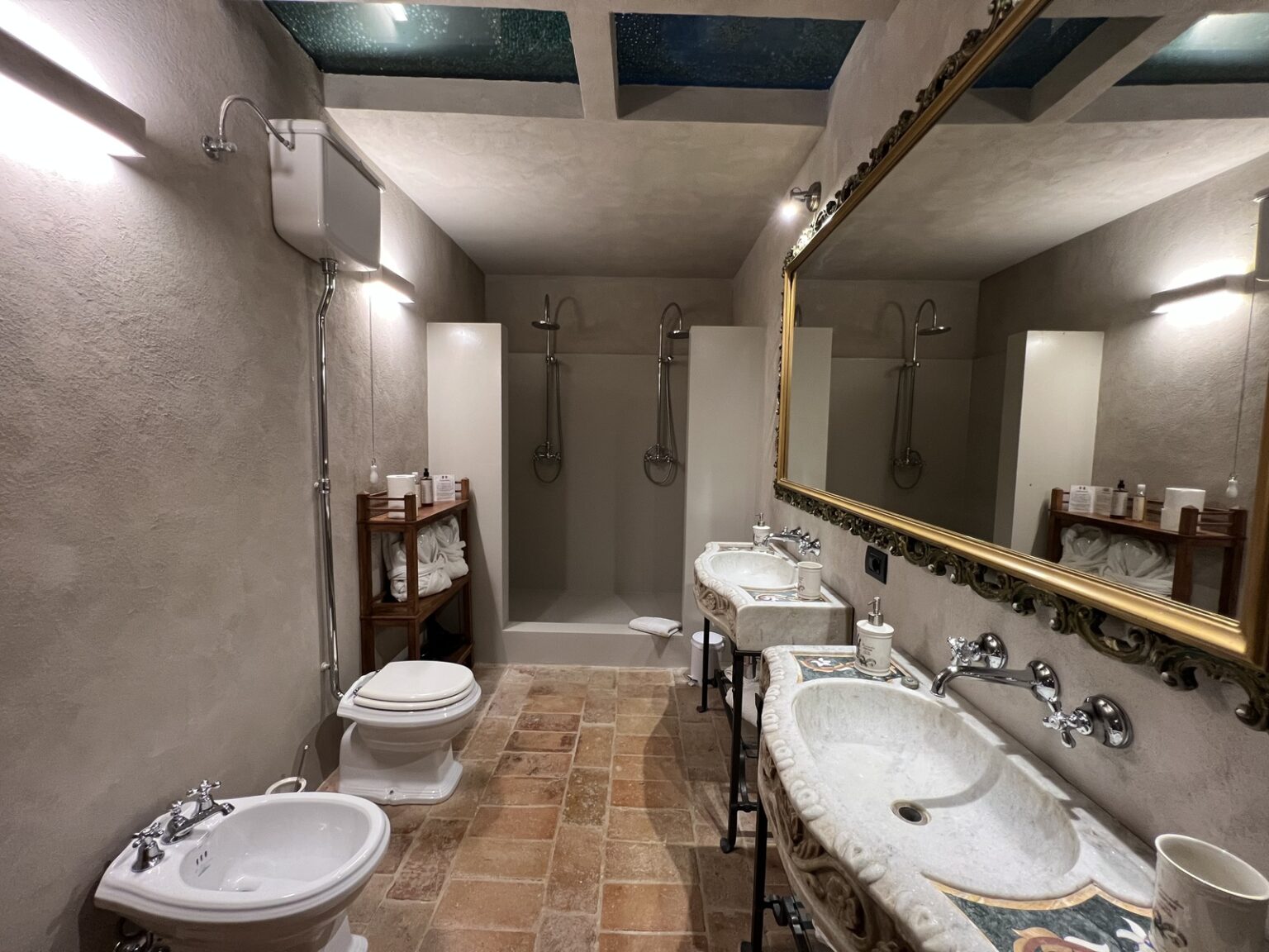 Spacious and elegantly designed bathroom featuring dual marble sinks, a modern shower area, a bidet, and a toilet. The room is illuminated by stylish wall lights and has a decorative mirror, complemented by natural stone flooring and soft color tones on the walls. | Only on SopranoVillas