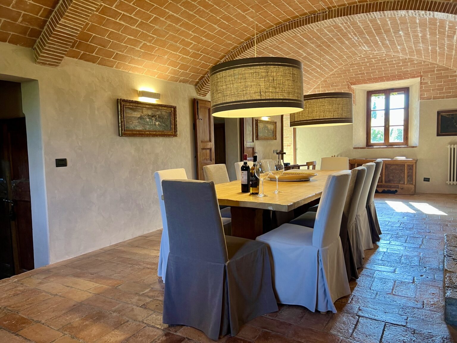 Spacious dining room featuring a wooden table surrounded by elegantly draped chairs, with a rustic brick ceiling and warm lighting. The room includes a wine display, glasses, and a scenic view through the windows, creating an inviting atmosphere for gatherings. | Only on SopranoVillas