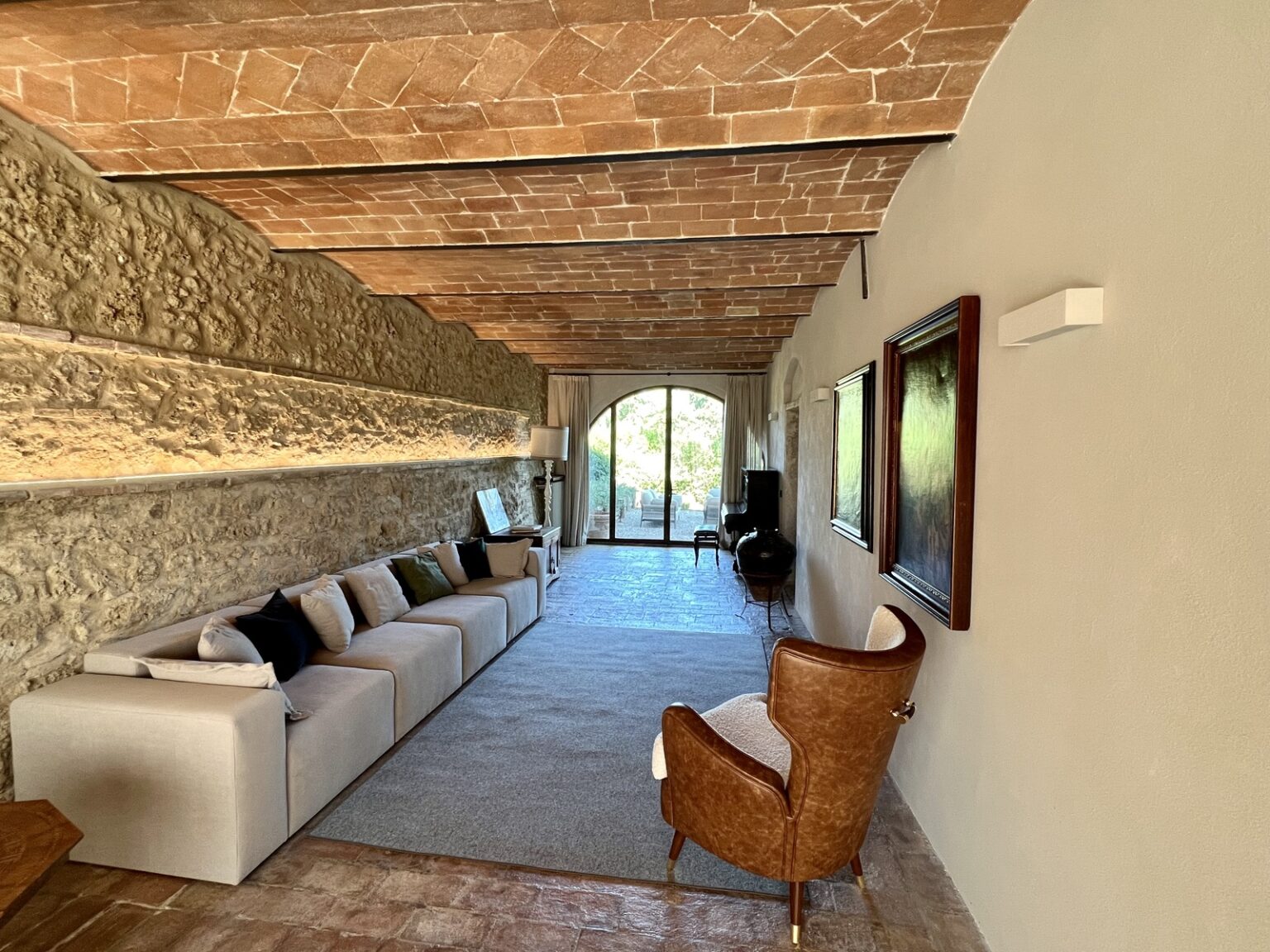 Modern living room interior featuring a beige sectional sofa with decorative pillows, exposed stone wall, and terracotta tile ceiling. The space is illuminated by soft lighting and includes a cozy brown armchair, wooden side table, and artwork on the walls, with large windows offering a view of the outdoors. | Only on SopranoVillas
