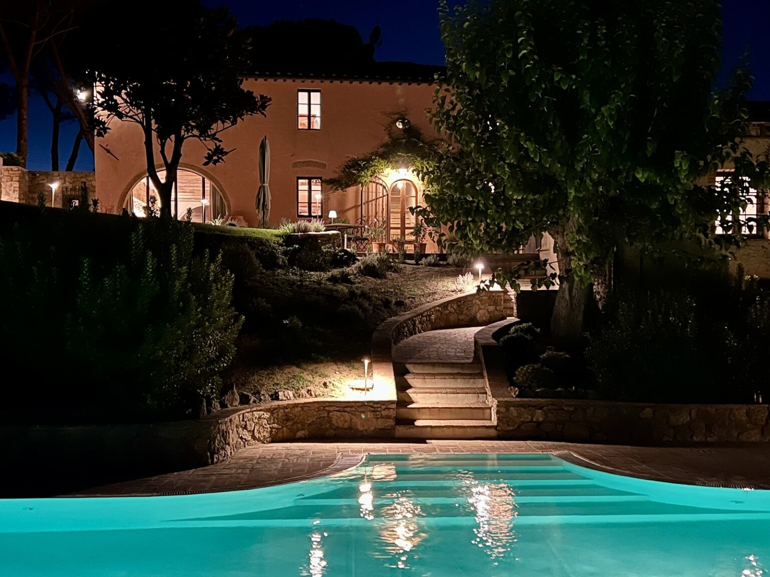 Illuminated outdoor pool area at night, featuring a stone pathway leading to a beautifully lit villa surrounded by lush greenery. | Only on SopranoVillas