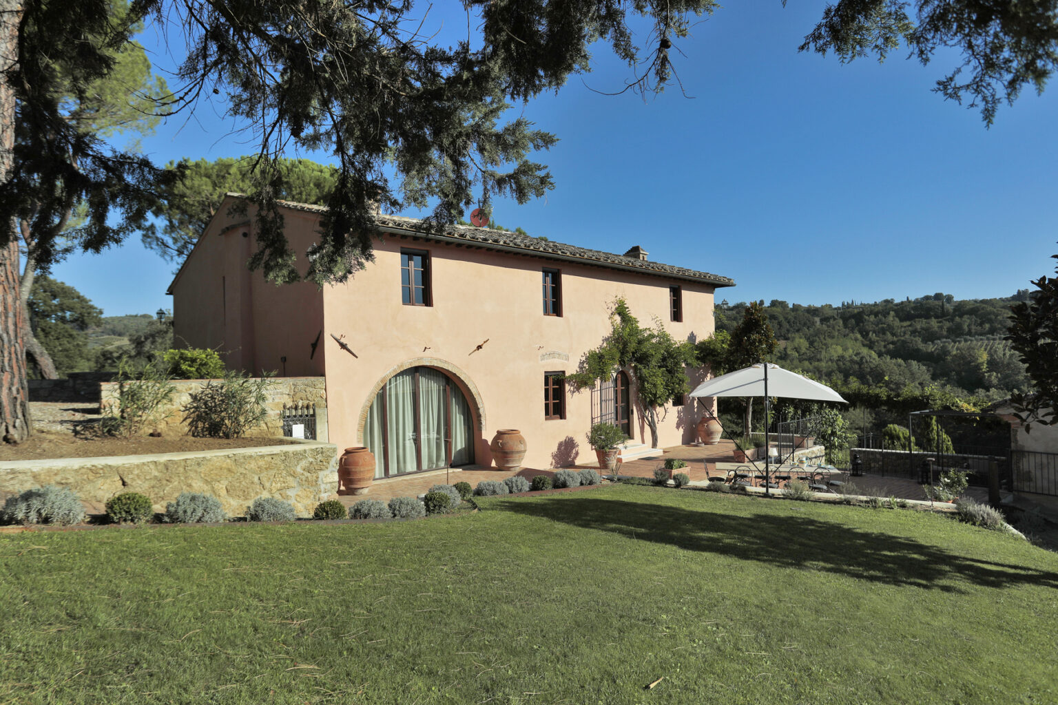 Charming pink villa surrounded by lush greenery, featuring a spacious lawn, outdoor seating area with an umbrella, and scenic views of the hillside. Ideal for a relaxing getaway in a serene setting. | Only on SopranoVillas