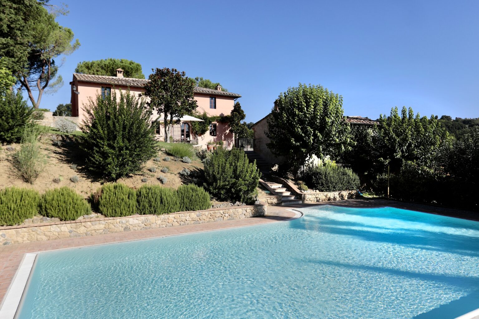 Scenic view of a Tuscan villa with a swimming pool surrounded by lush greenery under a clear blue sky. | Only on SopranoVillas