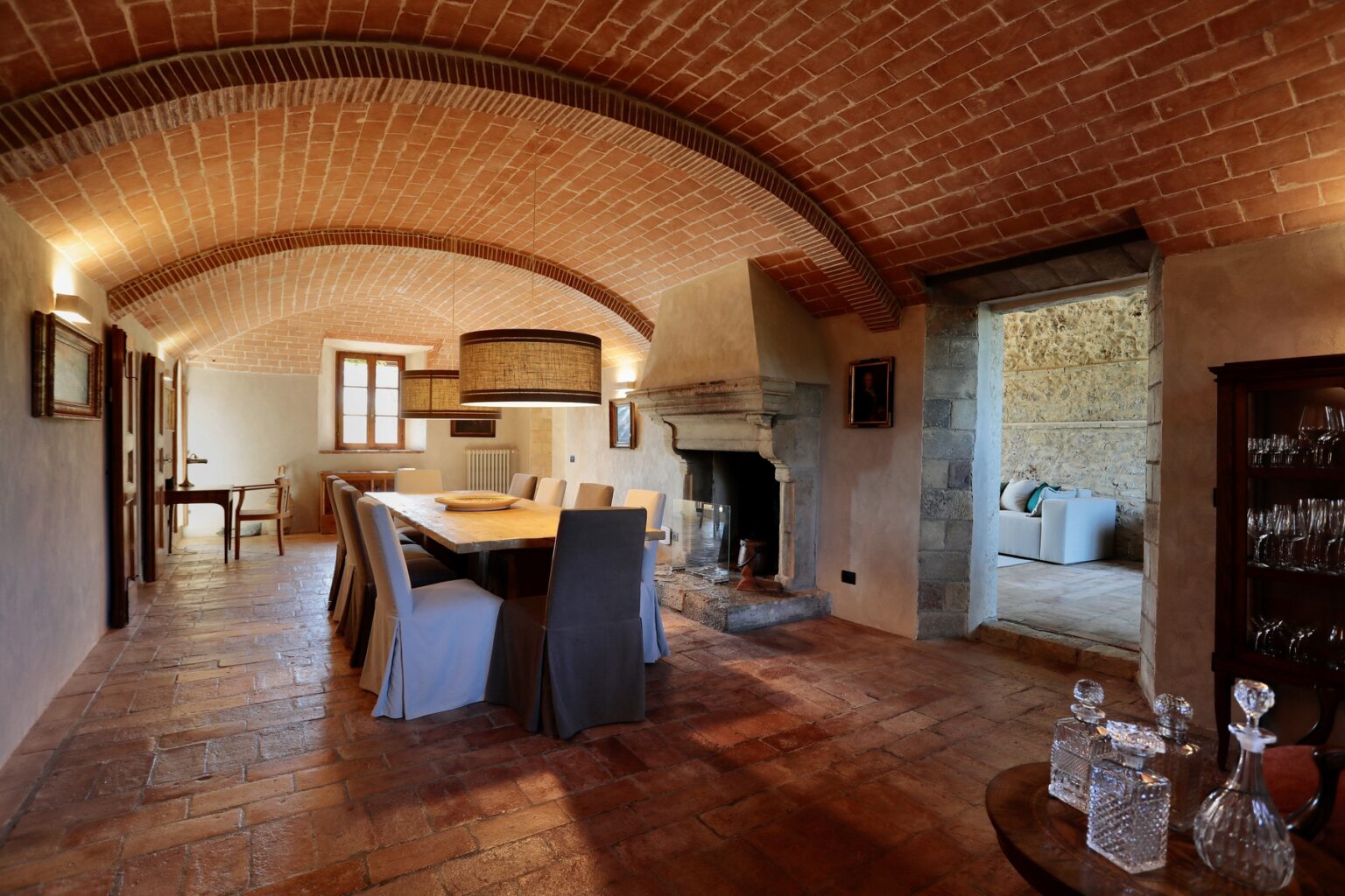Interior view of a rustic dining room featuring arched brick ceilings, a wooden dining table surrounded by upholstered chairs, and a cozy stone fireplace. Natural light streams in through windows, highlighting the warm terracotta flooring and inviting decor, with a glimpse of a modern living area in the background. | Only on SopranoVillas