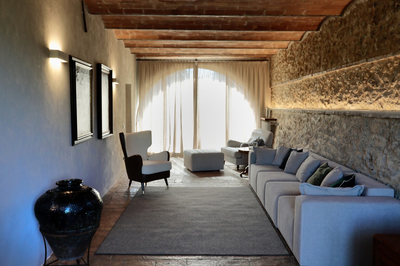 Modern living room interior featuring a comfortable white sofa, stylish armchair, and natural stone wall accents, illuminated by soft natural light from large windows. | Only on SopranoVillas
