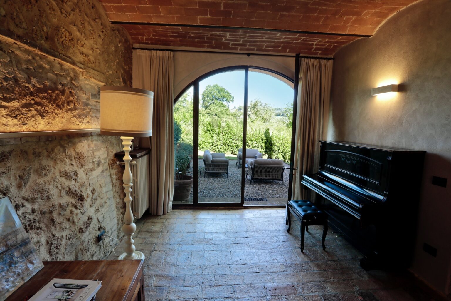 Cozy interior of a rustic room featuring a grand piano, stylish floor lamp, and large glass doors that open to a lush garden view. The room showcases natural stone walls, terracotta ceiling, and wooden furniture, creating a warm and inviting atmosphere. | Only on SopranoVillas