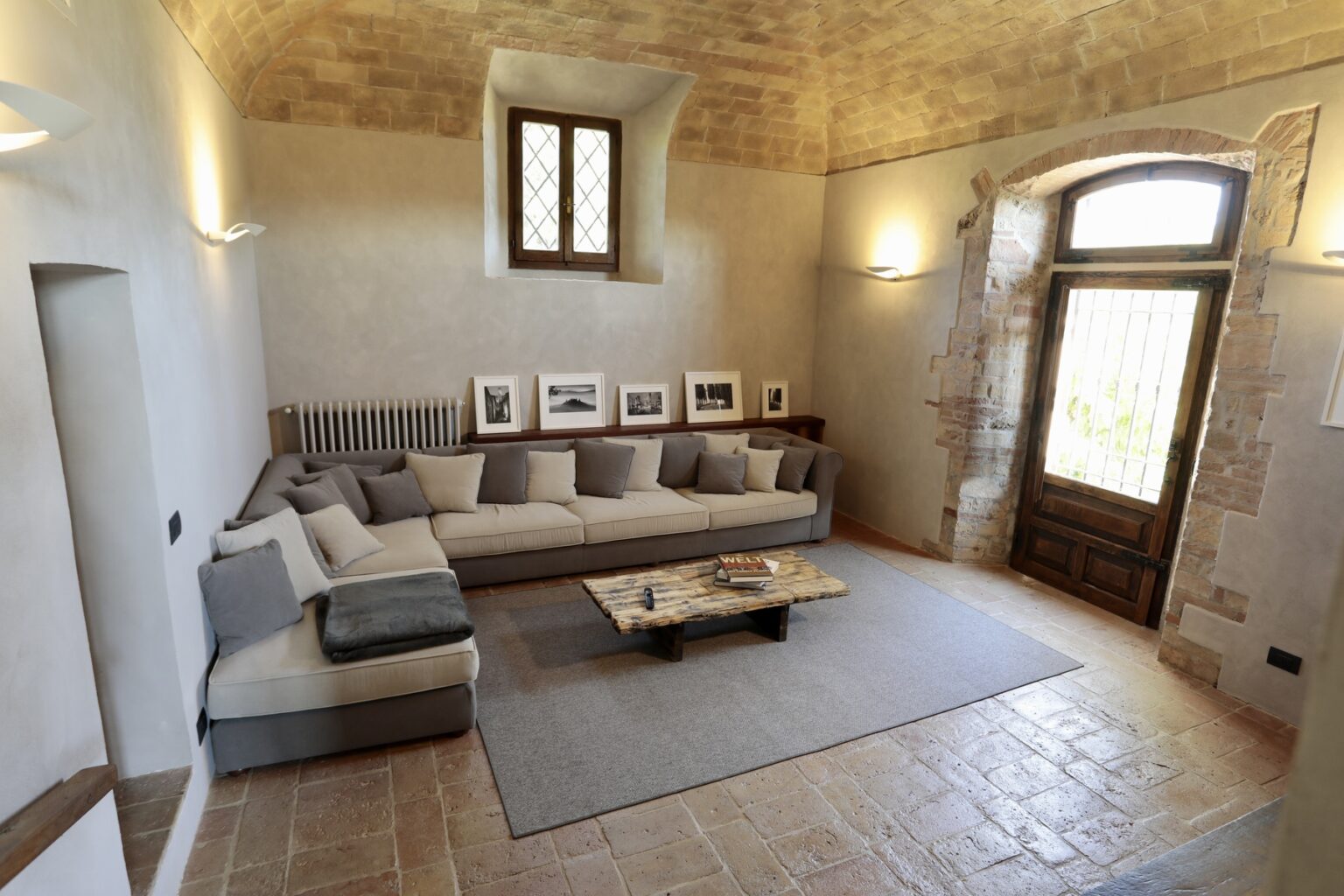 Spacious living room featuring a comfortable gray sectional sofa, a wooden coffee table, and natural light streaming through large windows, with stone walls and rustic decor elements. | Only on SopranoVillas